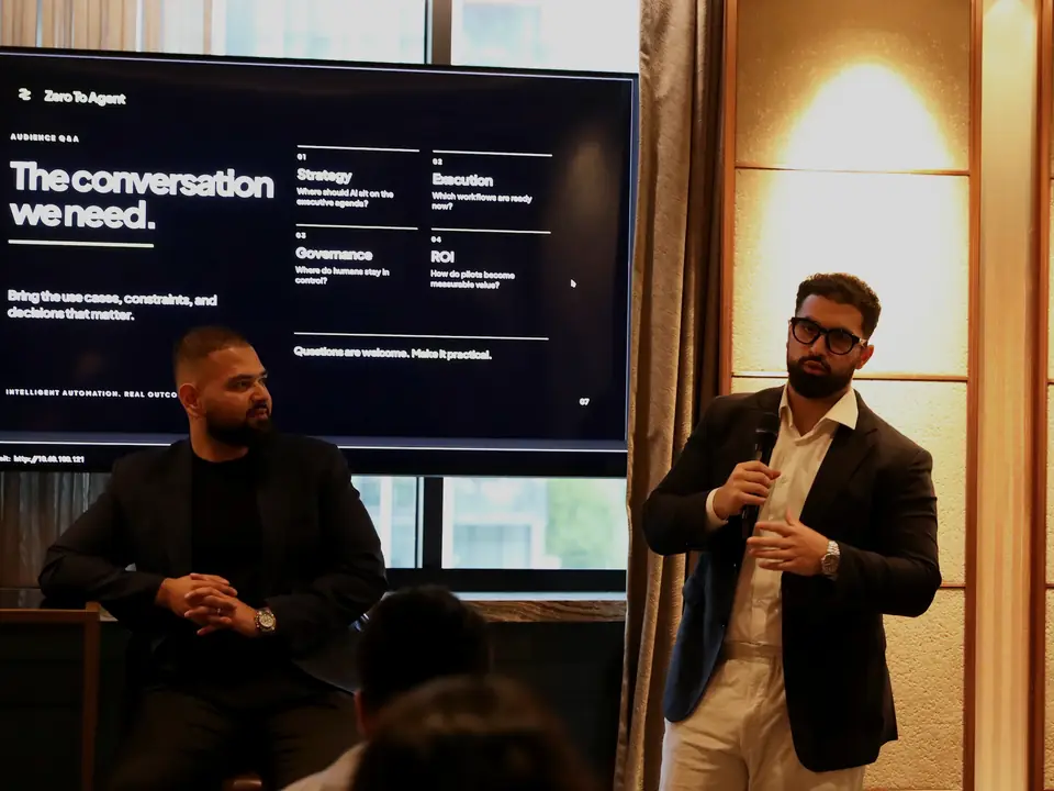 A team member speaking into a microphone at a Zero to Agent event, beside a screen that reads The conversation we need.